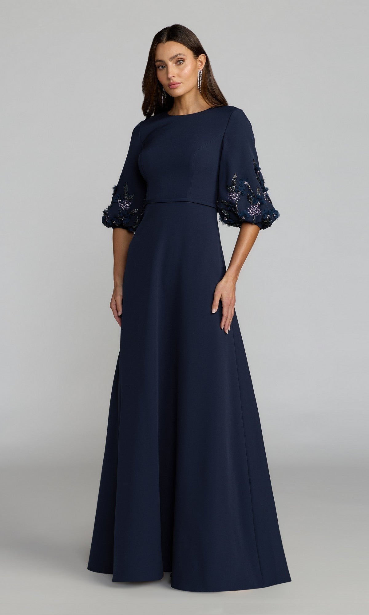 A woman wearing the Mac Duggal 13230 Long Formal Dress in Midnight Multi, featuring three-quarter sleeves adorned with floral embellishments, stands against a plain light gray background.